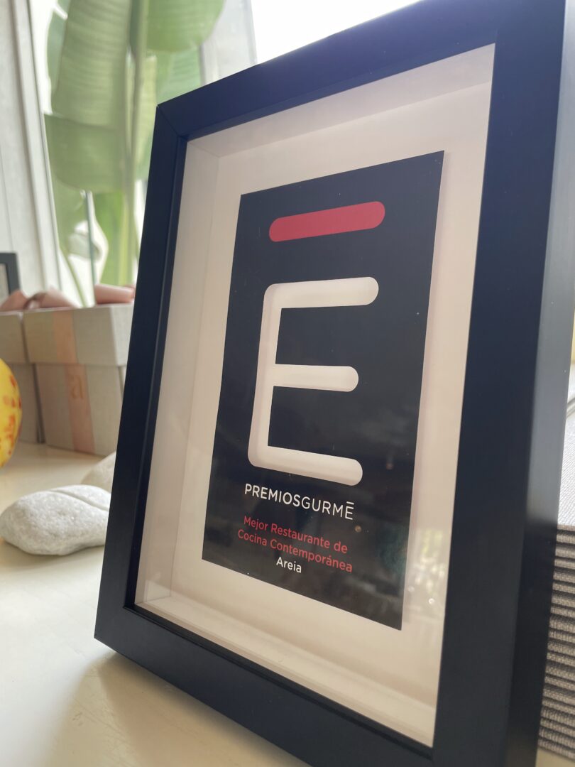 Framed Premios Gurmé Málaga plaque awarding Areia Best Contemporary Cuisine Restaurant, next to a plant and white stones.